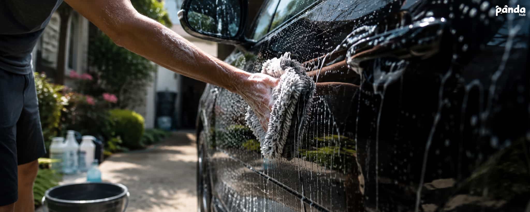 Person hand washing the side of a black car with a microfiber wash mitt, covered in soap suds in a residential driveway.