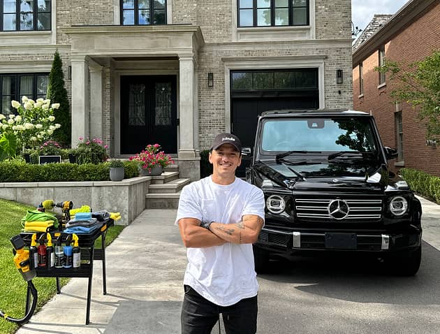 A professional car detailer in white t-shirt and black Panda Hub cap smiling with arms crossed in front of a black Mercedes-Benz G-Class SUV, with car detailing supplies arranged on a table in a residential driveway.