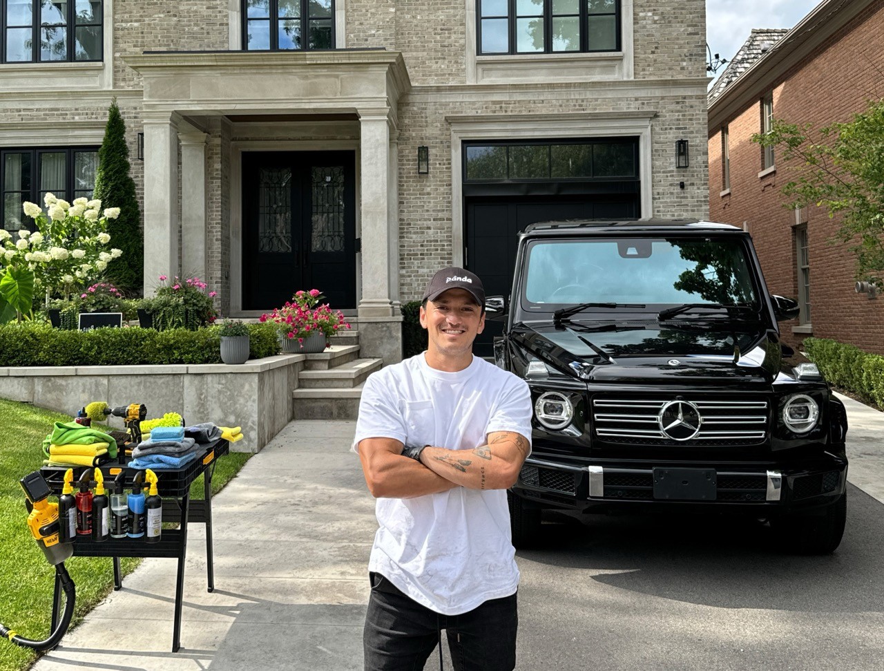 A professional car detailer in white t-shirt and black Panda Hub cap smiling with arms crossed in front of a black Mercedes-Benz G-Class SUV, with car detailing supplies arranged on a table in a residential driveway.